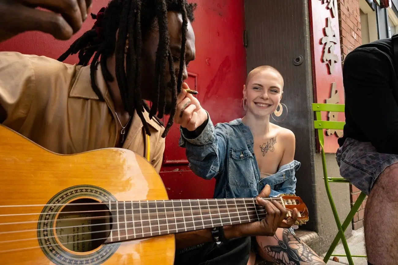 Friends sharing a joint in the street, with one woman holding the final tokes up to the mouth of a man playing guitar.